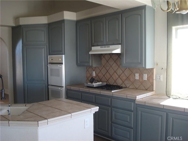 kitchen with tile counters, tasteful backsplash, stainless steel gas cooktop, white double oven, and under cabinet range hood