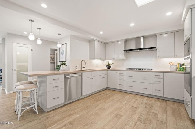 kitchen featuring light wood-style flooring, a peninsula, wall chimney range hood, appliances with stainless steel finishes, and a sink