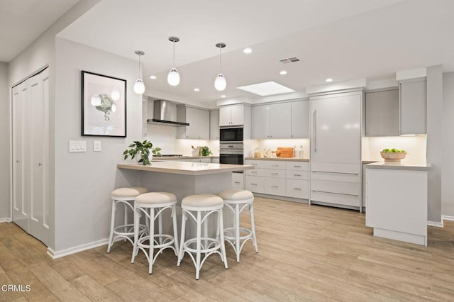 kitchen featuring a peninsula, light wood-type flooring, wall chimney range hood, visible vents, and appliances with stainless steel finishes