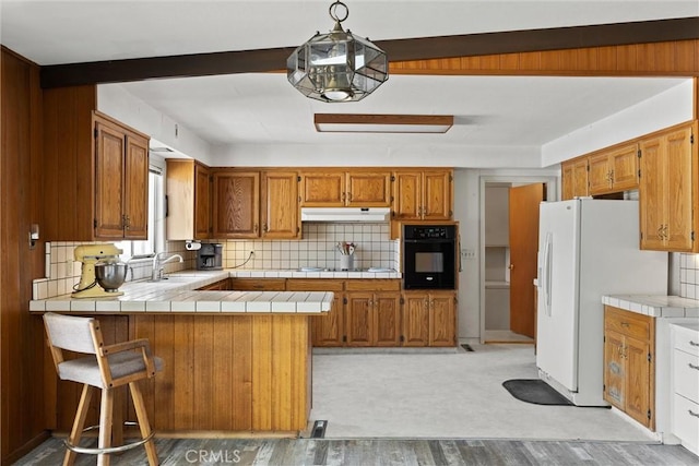kitchen featuring a sink, oven, white fridge with ice dispenser, a peninsula, and under cabinet range hood
