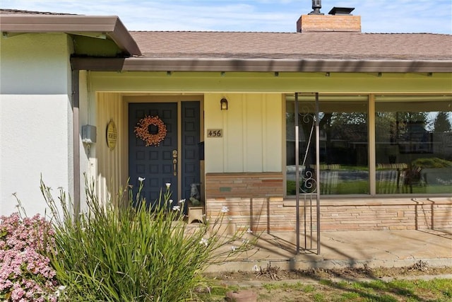 view of exterior entry with board and batten siding, a shingled roof, a chimney, and covered porch