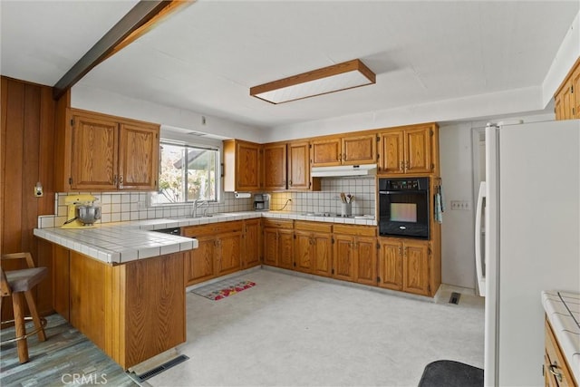 kitchen with visible vents, freestanding refrigerator, brown cabinetry, under cabinet range hood, and black oven