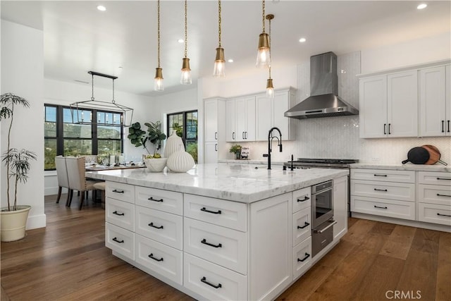 kitchen with dark wood-style flooring, a kitchen island with sink, tasteful backsplash, pendant lighting, and wall chimney range hood