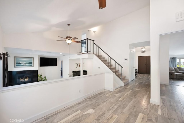 unfurnished living room with wood finished floors, stairs, a glass covered fireplace, high vaulted ceiling, and a ceiling fan