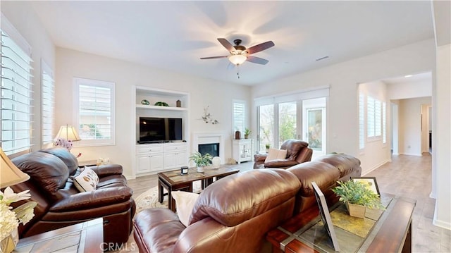 living area with a glass covered fireplace, light wood-style floors, visible vents, a ceiling fan, and built in features