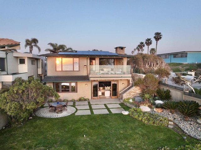 back of property with stucco siding, solar panels, a patio area, a balcony, and a lawn