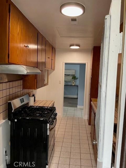 kitchen with brown cabinets, under cabinet range hood, stainless steel gas range, and light tile patterned floors
