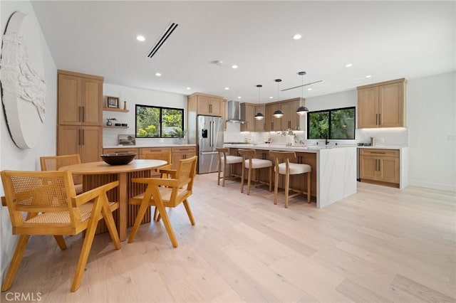 kitchen with wall chimney range hood, visible vents, stainless steel refrigerator with ice dispenser, a center island, and light wood finished floors