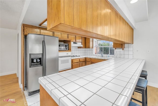 kitchen with tasteful backsplash, stainless steel appliances, under cabinet range hood, tile countertops, and a peninsula