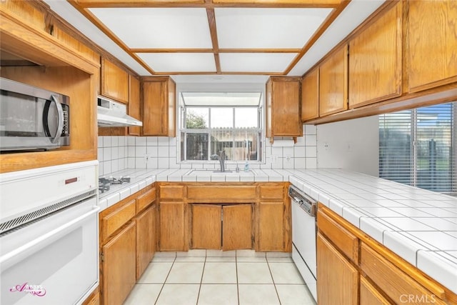 kitchen with white appliances, light tile patterned floors, under cabinet range hood, backsplash, and a sink