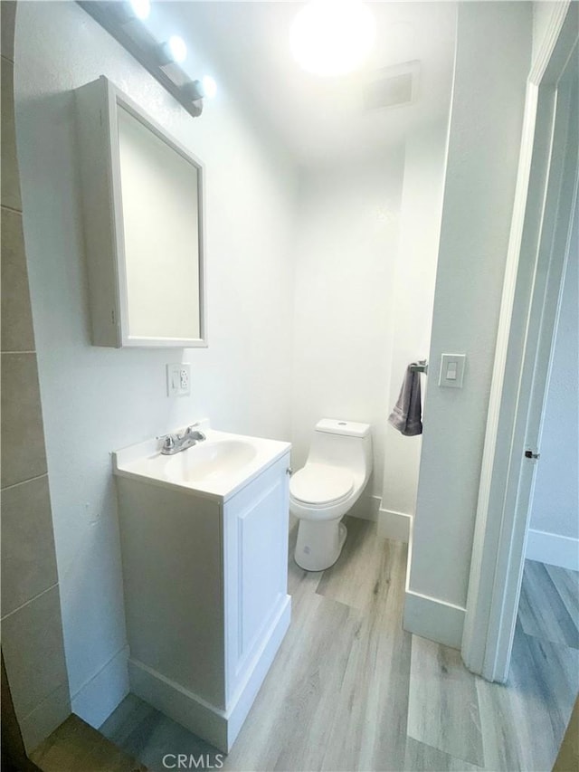 half bathroom with toilet, vanity, wood finished floors, baseboards, and visible vents
