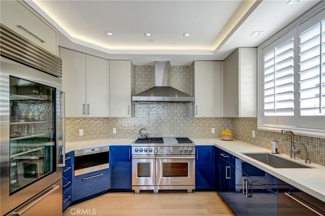 kitchen with blue cabinetry, light countertops, a sink, high end appliances, and wall chimney exhaust hood