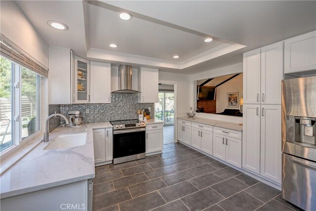 kitchen featuring a sink, a raised ceiling, stainless steel appliances, wall chimney range hood, and decorative backsplash