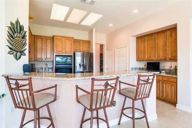 kitchen featuring a kitchen bar, black appliances, light stone counters, a peninsula, and brown cabinetry