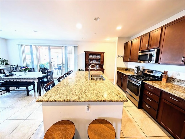 kitchen featuring a sink, light tile patterned floors, a center island with sink, light stone counters, and appliances with stainless steel finishes