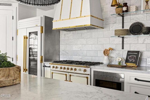 kitchen with backsplash, built in appliances, wall chimney range hood, white cabinets, and light stone countertops