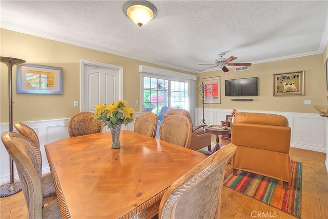dining room with a wainscoted wall, a textured ceiling, ceiling fan, ornamental molding, and light wood-type flooring