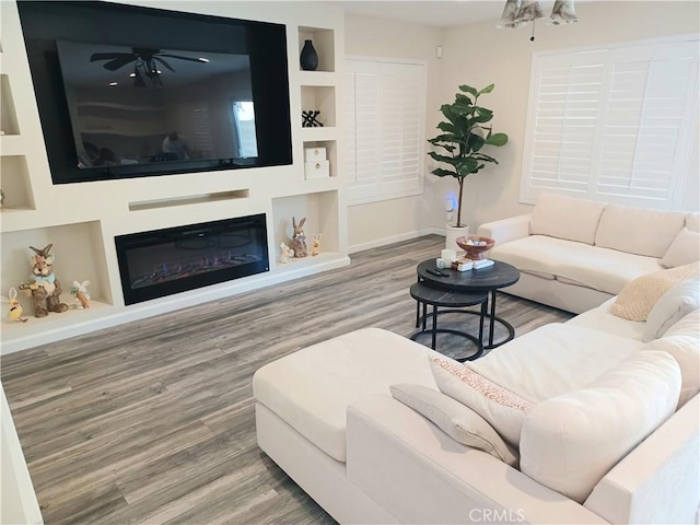 living room featuring wood finished floors, built in features, a glass covered fireplace, and a ceiling fan