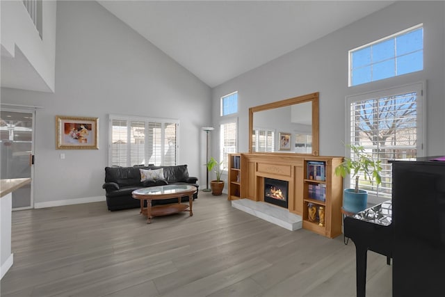 living room featuring baseboards, high vaulted ceiling, a lit fireplace, and light wood finished floors