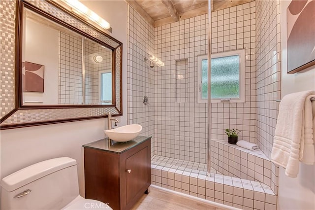 bathroom with vanity, beamed ceiling, toilet, and tiled shower