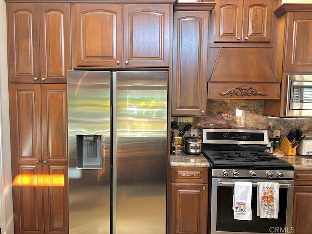 kitchen with stainless steel appliances, brown cabinetry, tasteful backsplash, light stone counters, and premium range hood