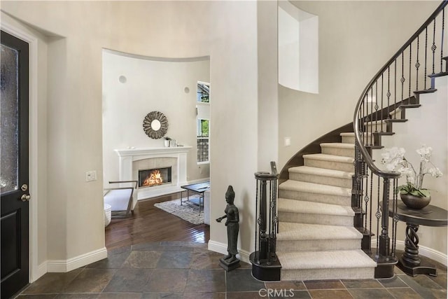 foyer entrance with stone finish flooring, stairway, a high ceiling, a glass covered fireplace, and baseboards