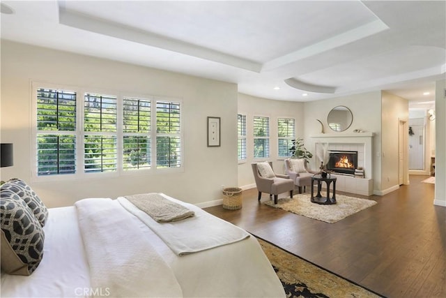 bedroom featuring a tray ceiling, a tiled fireplace, dark wood finished floors, baseboards, and recessed lighting