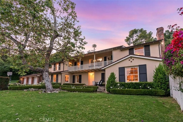 back of property at dusk with a lawn, stucco siding, a balcony, and a chimney