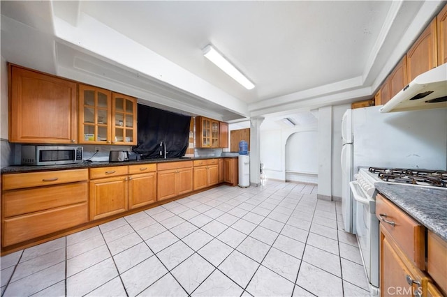 kitchen featuring white range with gas stovetop, brown cabinetry, light tile patterned floors, glass insert cabinets, and stainless steel microwave