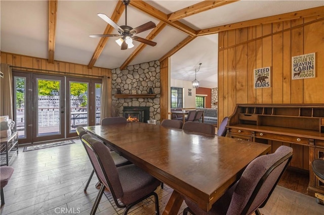 dining room featuring wooden walls, beam ceiling, a stone fireplace, a ceiling fan, and hardwood / wood-style flooring