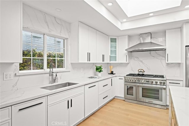 kitchen with range with two ovens, a sink, light wood finished floors, wall chimney range hood, and white cabinets