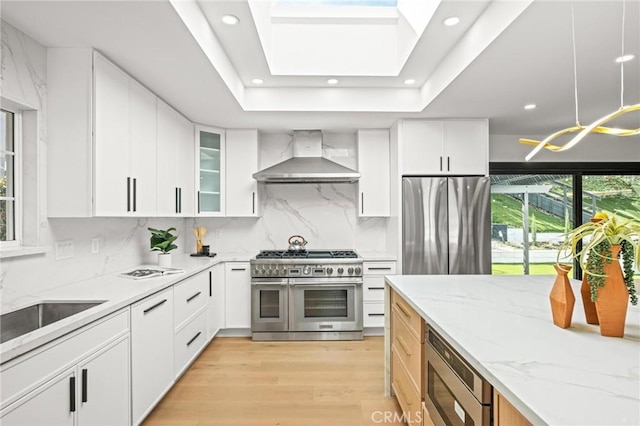 kitchen featuring tasteful backsplash, light stone counters, light wood finished floors, wall chimney range hood, and appliances with stainless steel finishes