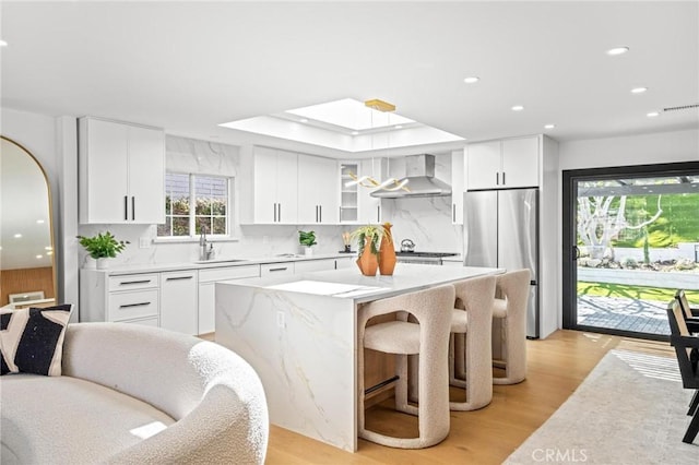 kitchen with a sink, freestanding refrigerator, a center island, light wood-type flooring, and wall chimney range hood