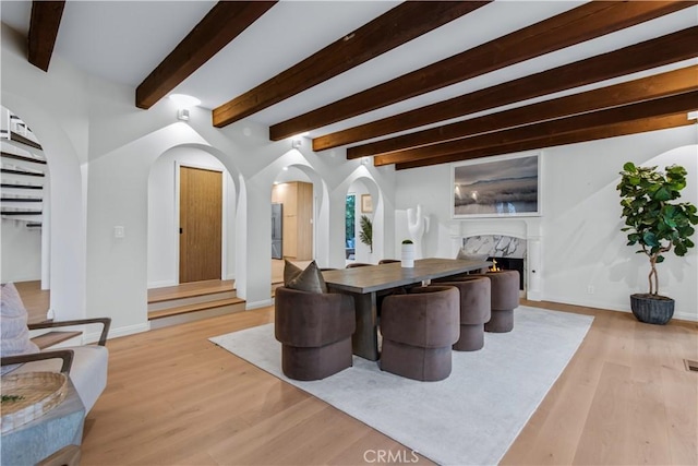 dining space with baseboards, a fireplace, beam ceiling, arched walkways, and light wood-style floors