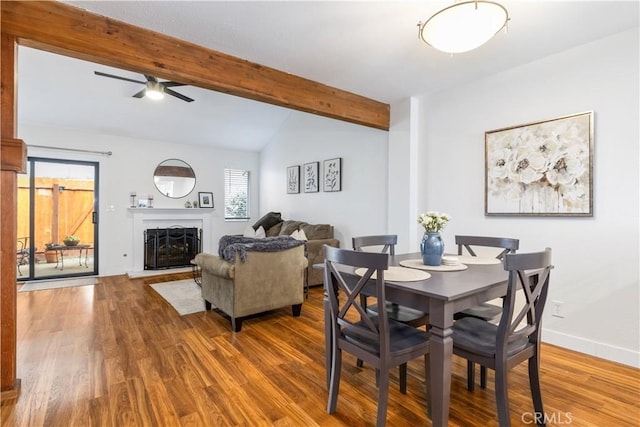dining space with a ceiling fan, a fireplace with raised hearth, wood finished floors, and vaulted ceiling with beams