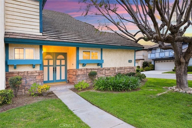 exterior entry at dusk featuring a yard, stucco siding, stone siding, a tile roof, and french doors