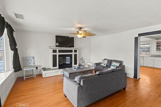 living room featuring visible vents, a ceiling fan, light wood-style floors, a textured ceiling, and a glass covered fireplace