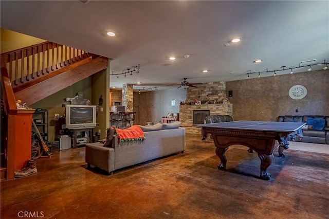 game room featuring a stone fireplace, rail lighting, ceiling fan, and concrete flooring