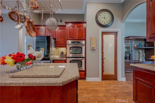 kitchen with stainless steel double oven, light wood-style floors, crown molding, arched walkways, and dark brown cabinets