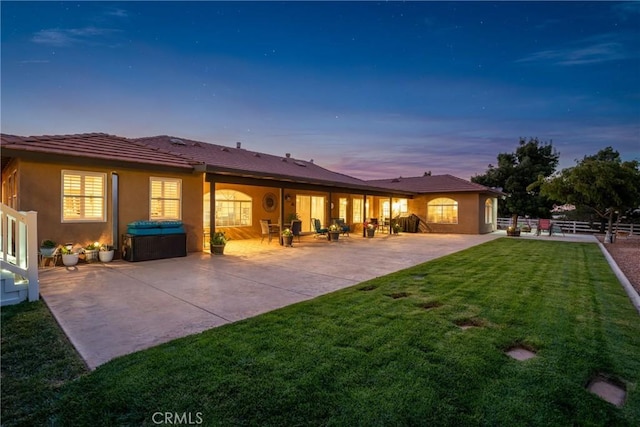 back of house at dusk featuring fence, a patio, a hot tub, stucco siding, and a yard