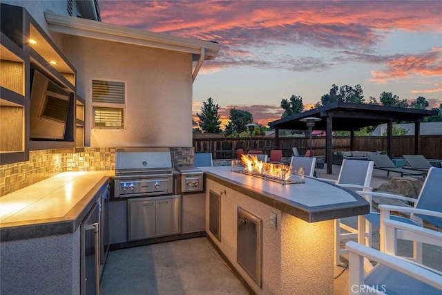 patio terrace at dusk featuring area for grilling, exterior kitchen, fence, and an outdoor fire pit