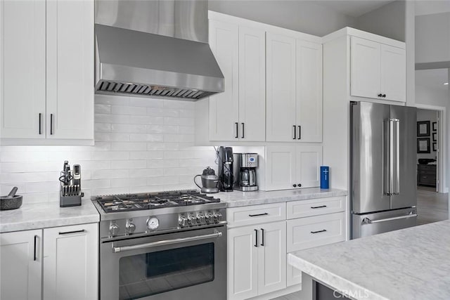 kitchen featuring high end appliances, decorative backsplash, wall chimney range hood, and white cabinetry