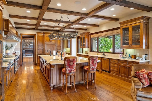 kitchen featuring dark wood finished floors, a sink, a kitchen island with sink, brown cabinetry, and glass insert cabinets