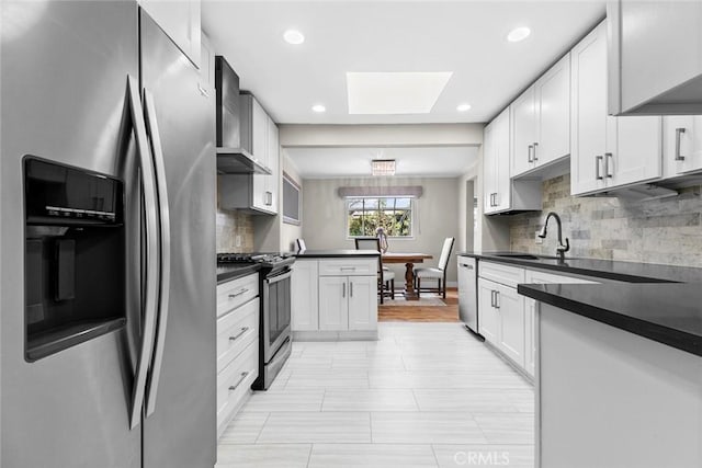 kitchen with a skylight, wall chimney exhaust hood, dark countertops, appliances with stainless steel finishes, and a sink