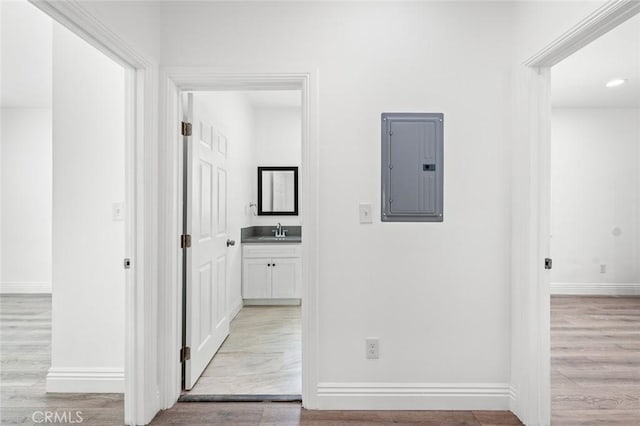 corridor featuring a sink, baseboards, light wood finished floors, and electric panel