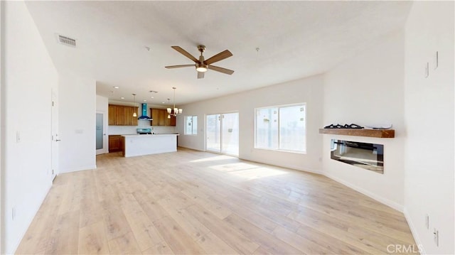 unfurnished living room with light wood finished floors, visible vents, baseboards, a glass covered fireplace, and ceiling fan with notable chandelier