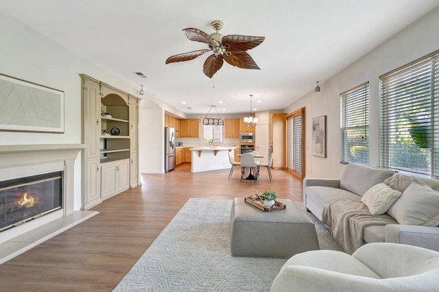 living area featuring ceiling fan with notable chandelier, visible vents, light wood-style flooring, and a glass covered fireplace