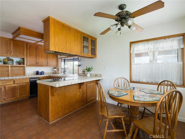 kitchen with a ceiling fan, tile counters, a peninsula, brown cabinets, and glass insert cabinets