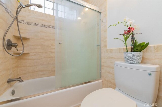 bathroom with tile walls, toilet, and shower / bath combination with glass door