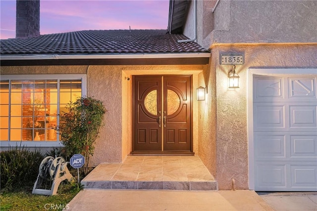 exterior entry at dusk with stucco siding, a chimney, a garage, and a tiled roof